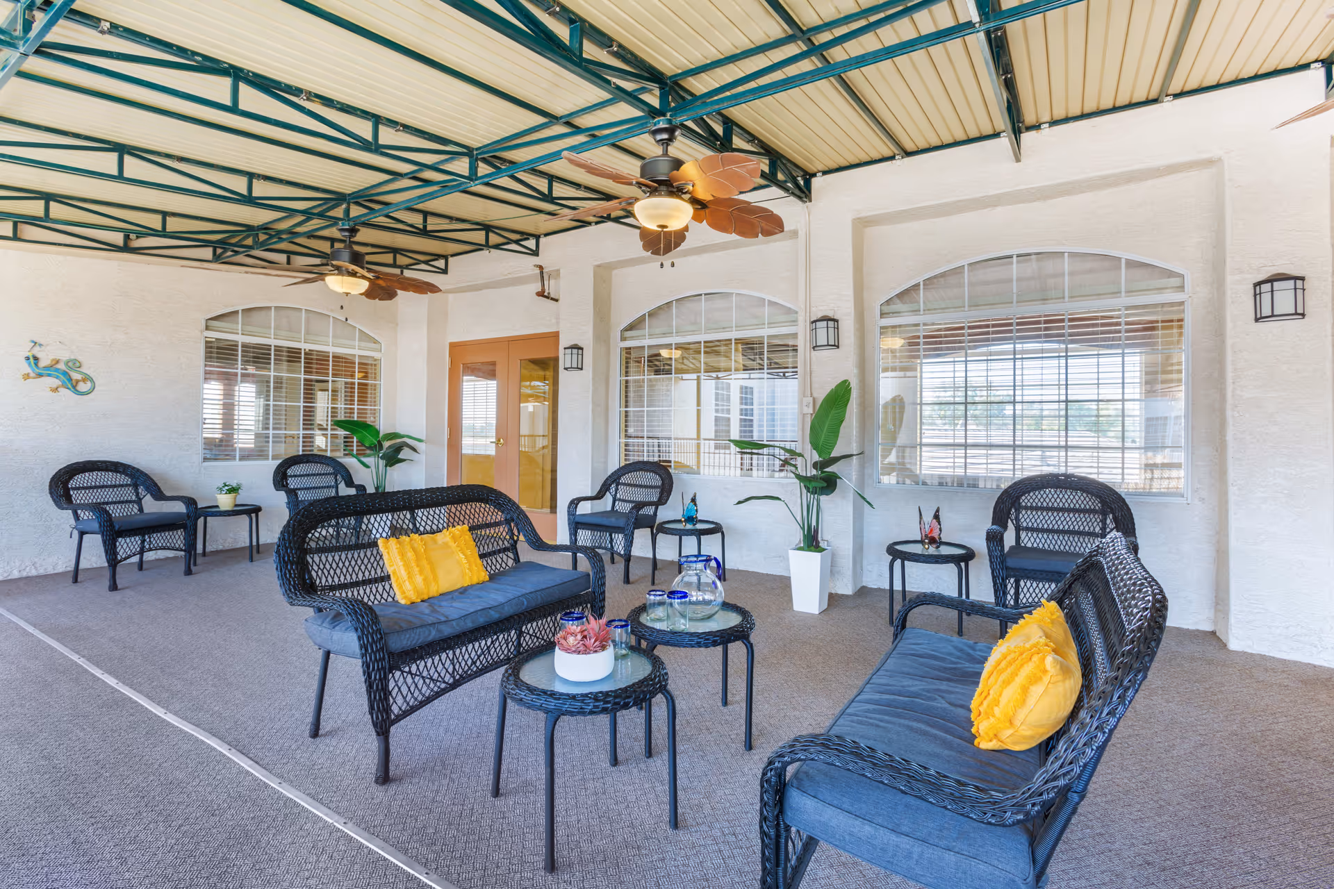 Covered outdoor seating area with black wicker furniture including two loveseats and several chairs, all with blue cushions and some with yellow pillows. There are small round tables with decorative items, ceiling fans with leaf-shaped blades, potted plants, and large windows with white blinds on the wall.