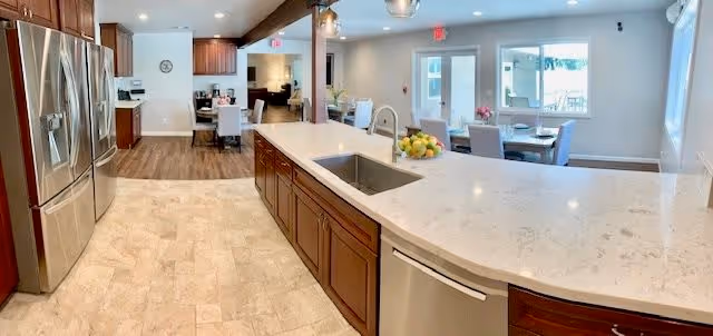 A spacious, modern kitchen area with a large marble countertop island featuring a built-in sink and dishwasher. Stainless steel refrigerator is on the left, and wooden cabinets line the walls. In the background, there is a dining area with tables and chairs near large windows letting in natural light.