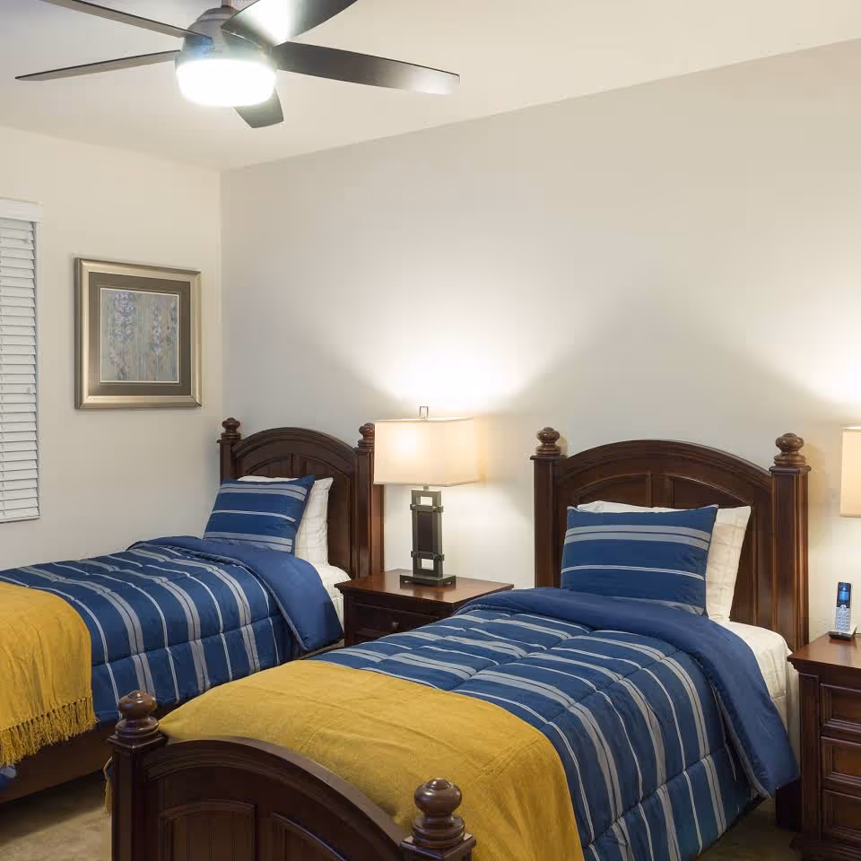 A neatly arranged bedroom with two twin beds featuring blue striped bedding and yellow throws, wooden headboards, bedside tables with lamps, and a ceiling fan.
