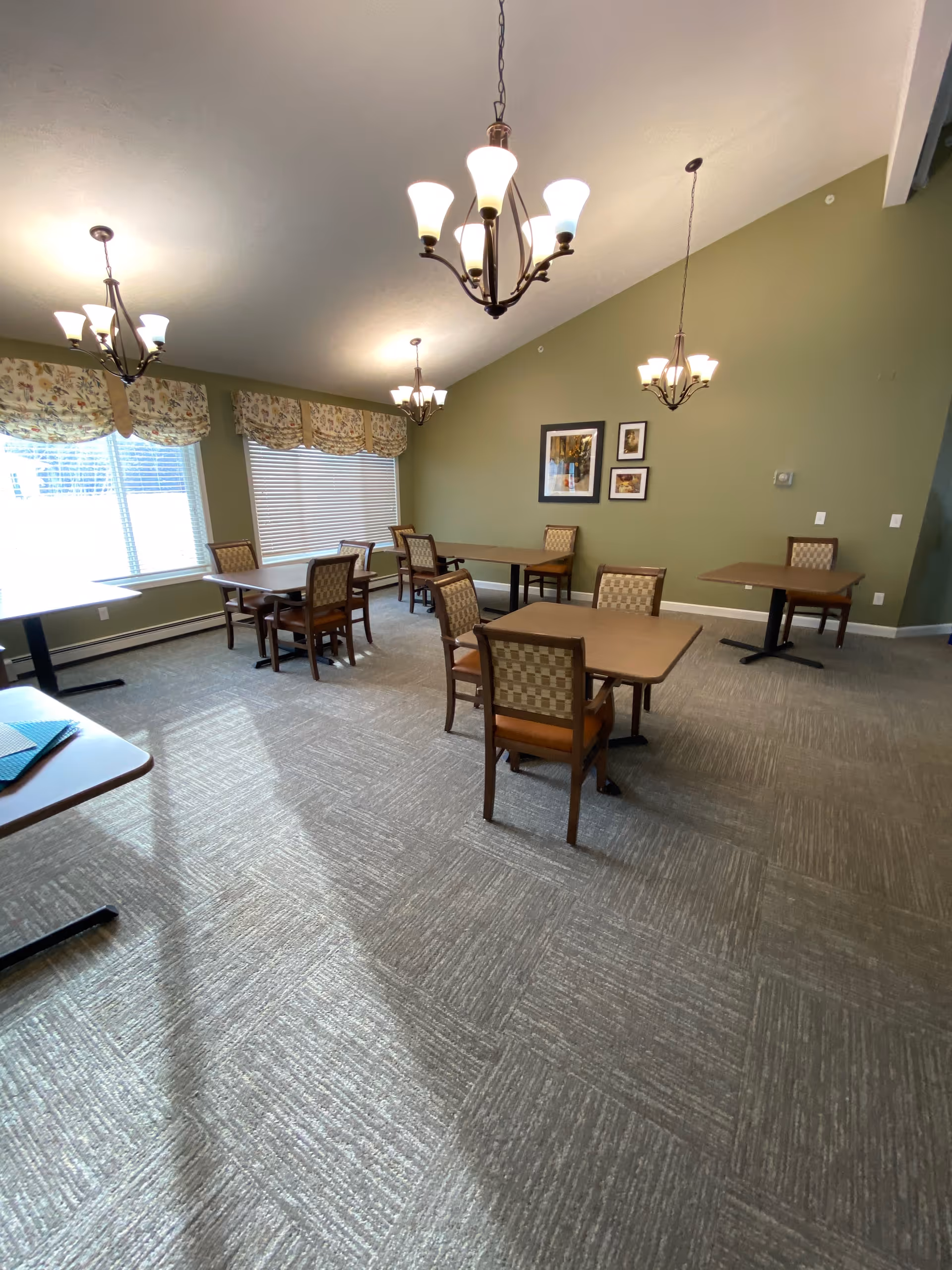 A dining room with several square tables and cushioned chairs arranged neatly on a carpeted floor. The walls are painted green and decorated with framed pictures. Large windows with floral valances allow natural light to enter the room. Multiple chandeliers hang from the ceiling providing additional lighting.