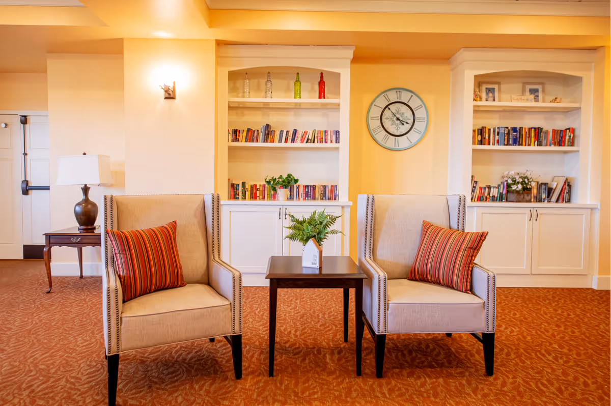 Two upholstered armchairs with striped pillows flank a small table in a cozy sitting area in front of built-in bookshelves and a wall clock.