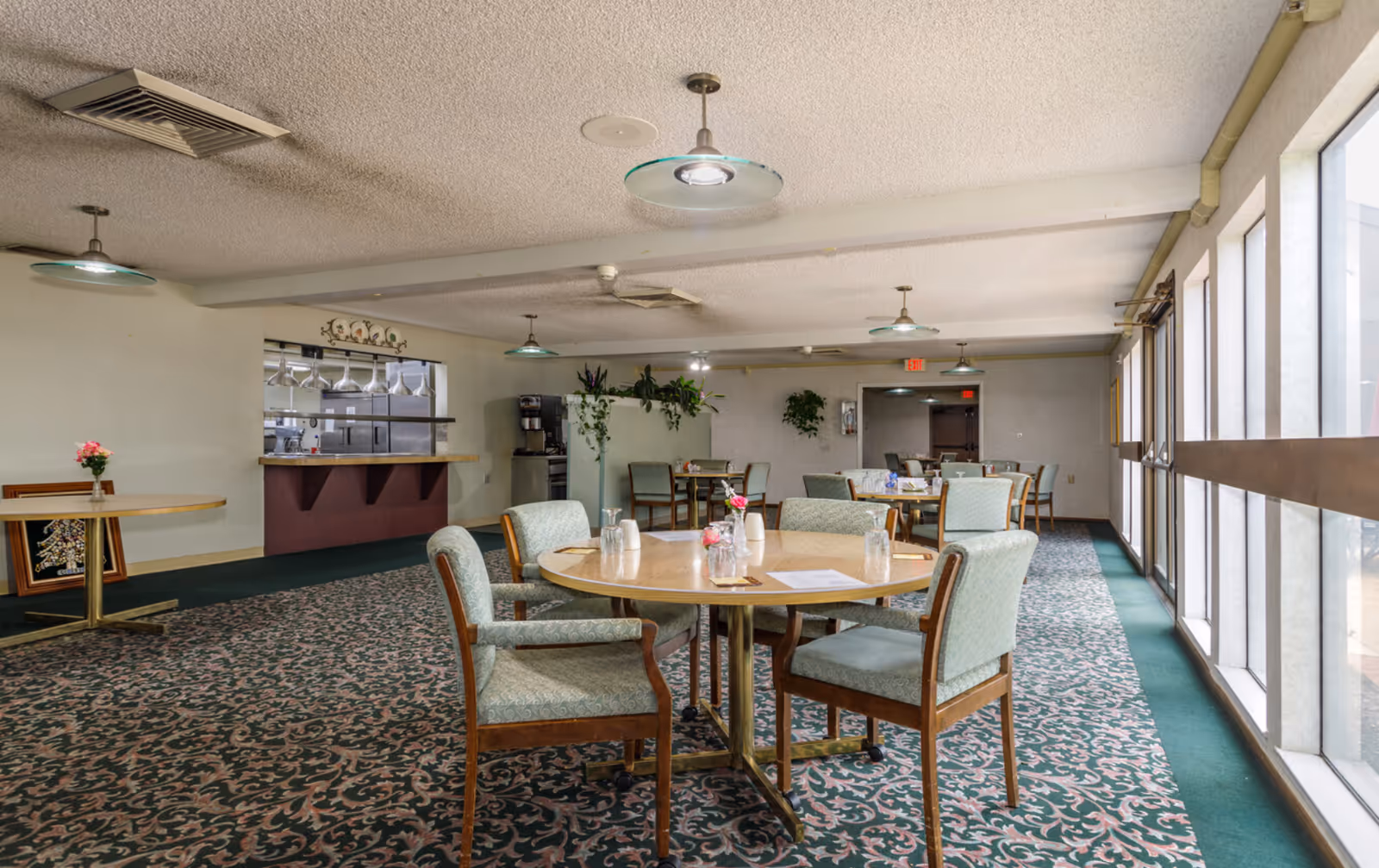Dining room with round tables and upholstered chairs, large windows along one side, and a serving counter in the background.
