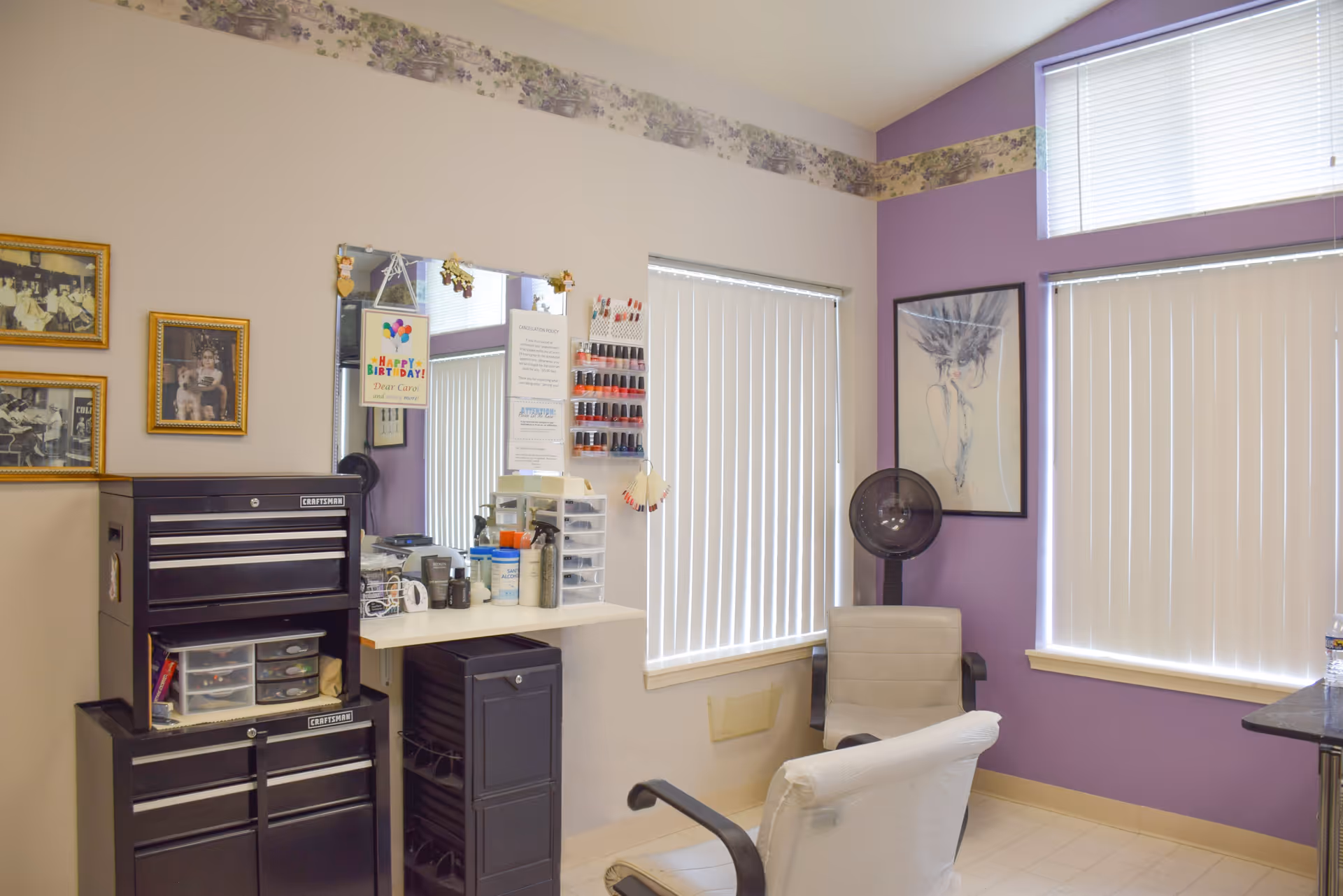 Interior of a small salon or beauty room with two chairs, a mirror on the wall, nail polish racks, a hair dryer, and various beauty supplies. The walls are painted white and purple with a floral border near the ceiling. There are two windows with vertical blinds partially closed.