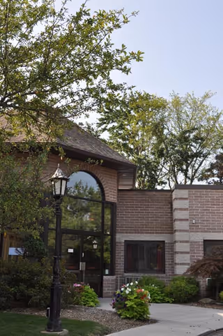 Exterior view of a nursing home building with brick walls, large arched glass windows, a black lamp post, and surrounding greenery including trees and flowering plants under a clear sky.