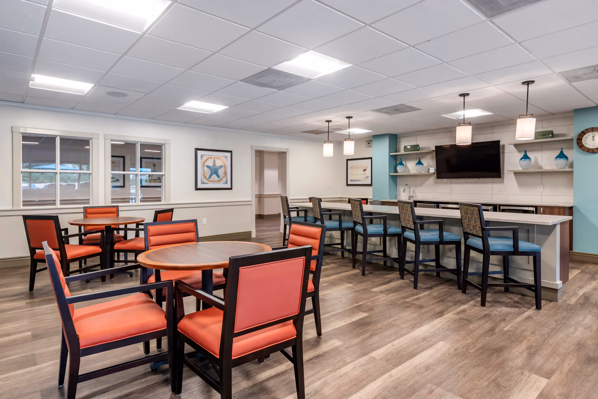 Bright communal dining/lounge area with round tables and red-upholstered chairs, a long counter with high stools, pendant lights, and a wall-mounted TV.