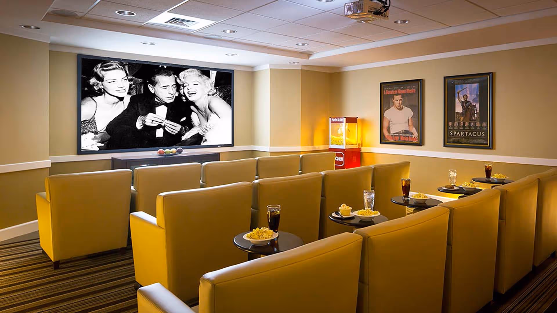 A cozy movie theater room with rows of yellow armchairs facing a large screen displaying a black and white movie scene. Small round tables in front of the chairs hold popcorn bowls and drinks. A popcorn machine is visible in the corner, and framed movie posters hang on the beige walls.