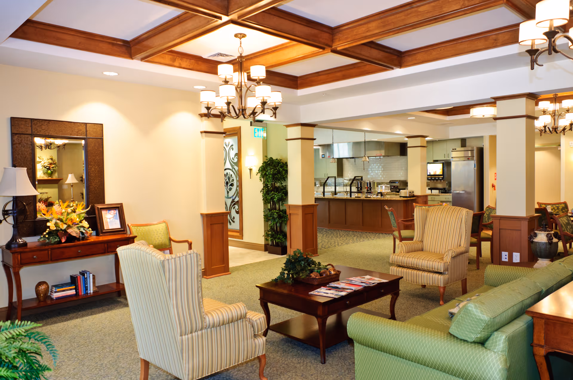 Bright, furnished communal lounge with armchairs, sofas, a coffee table, and a view into a kitchenette/dining area.