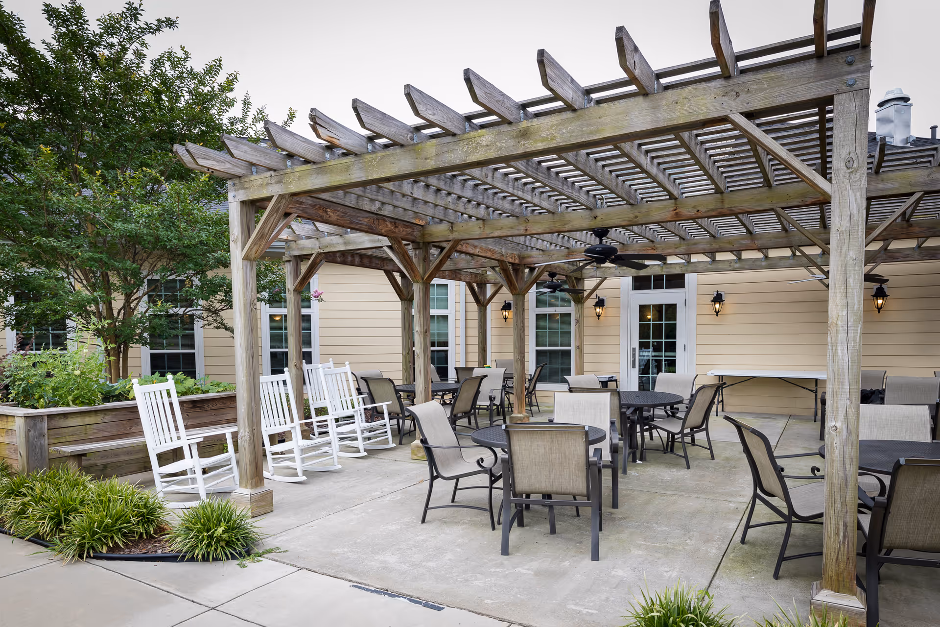 Outdoor patio area with a wooden pergola providing partial shade. The space includes several round tables with chairs and a row of white rocking chairs along a raised garden bed. The patio is adjacent to a building with beige siding and multiple windows and doors. There are ceiling fans mounted under the pergola and some wall-mounted lantern-style lights on the building exterior.