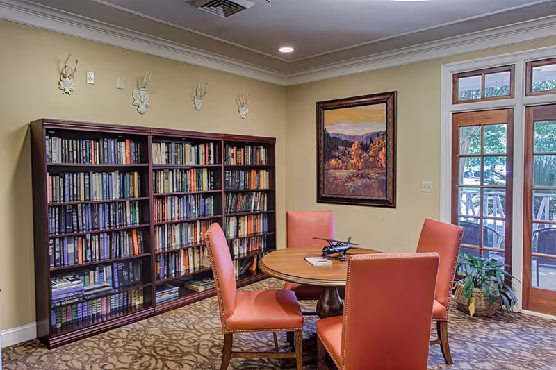 A cozy interior room featuring a round wooden table surrounded by four coral-colored upholstered chairs. Behind the table is a large bookshelf filled with books. On the wall above the bookshelf are four decorative white antler mounts. To the right, there is a framed landscape painting and a glass door leading to an outdoor area with greenery visible outside. A potted plant is placed near the door on the carpeted floor.