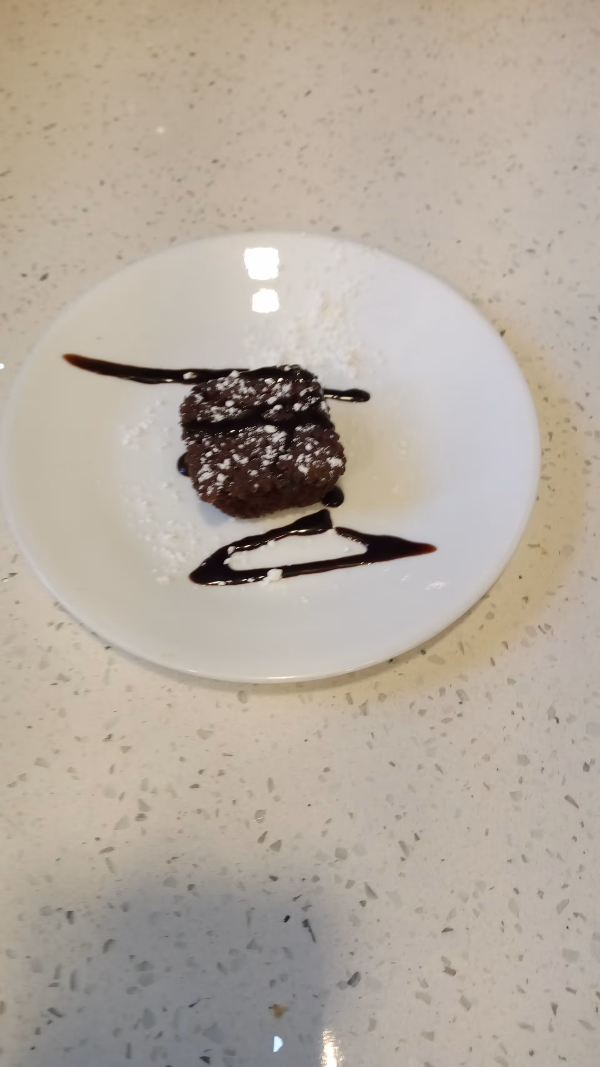 A small chocolate brownie dusted with powdered sugar on a white plate, decorated with chocolate syrup lines on a speckled countertop.