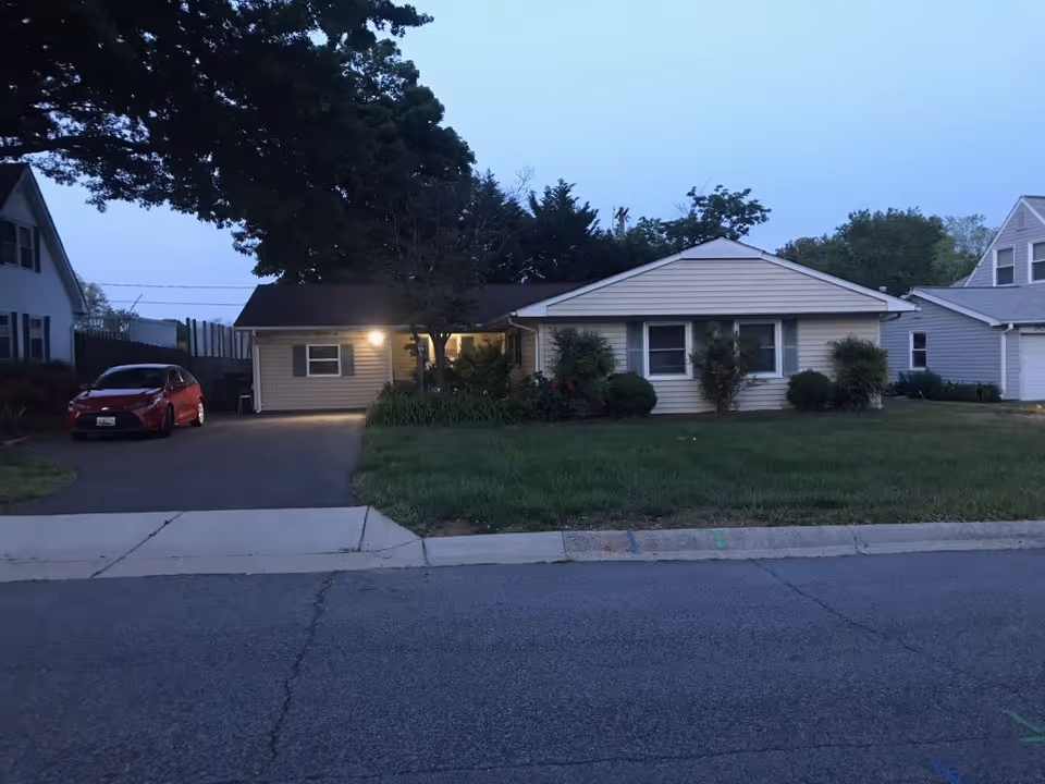Exterior view of a single-story residential building with beige siding and a dark roof, surrounded by grass and bushes. A driveway with a red car is visible on the left side, and the scene is captured during dusk with a light on near the entrance.