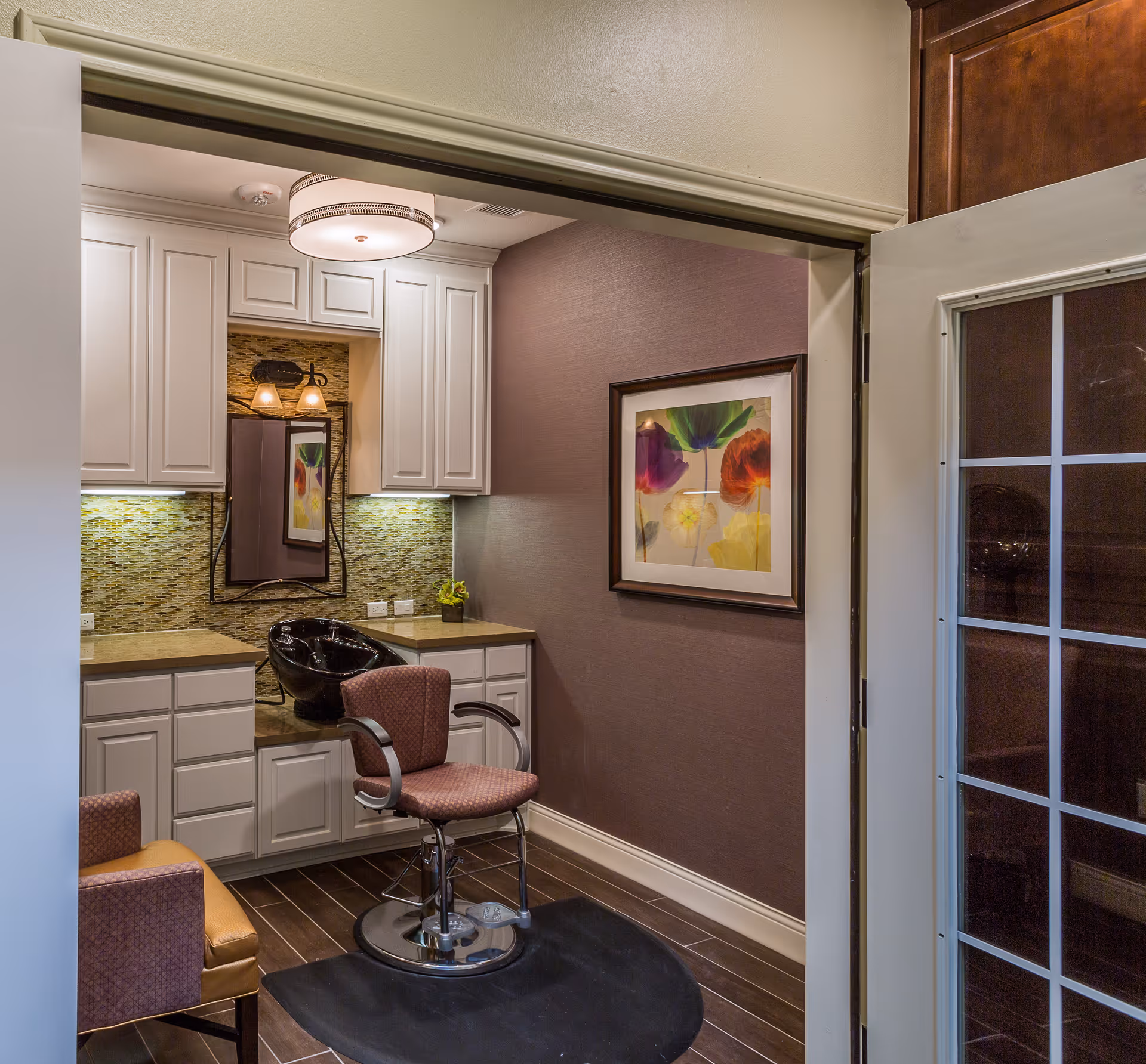 Interior view of a hair salon area in a senior living facility with a salon chair in front of a black wash basin, white cabinets, a mirror, and a framed colorful floral artwork on the wall.