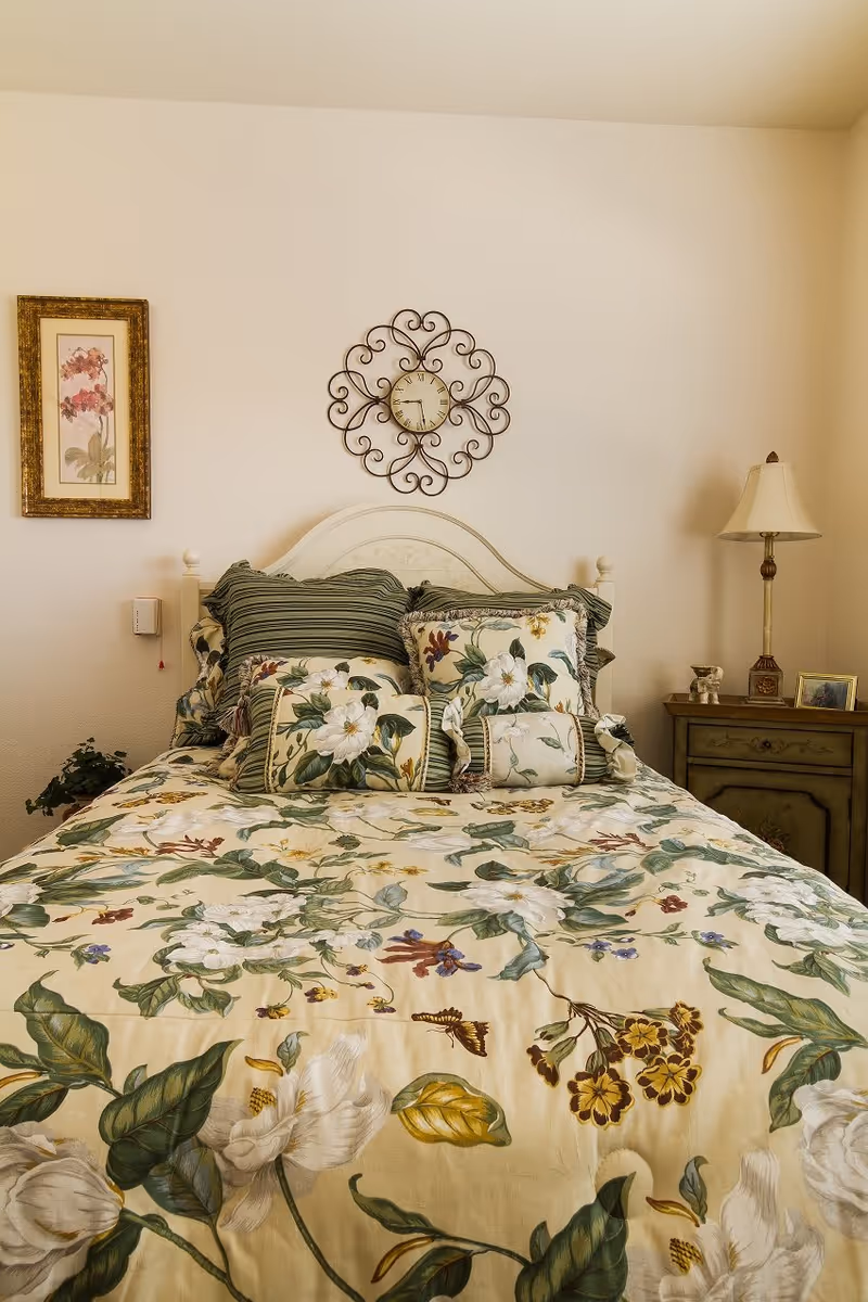 A bedroom with a bed covered in a floral patterned bedspread and matching pillows. Above the bed is a decorative wall clock with a wrought iron design. To the right of the bed is a wooden nightstand with a lamp, a small framed picture, and decorative items. On the left wall is a framed floral artwork.