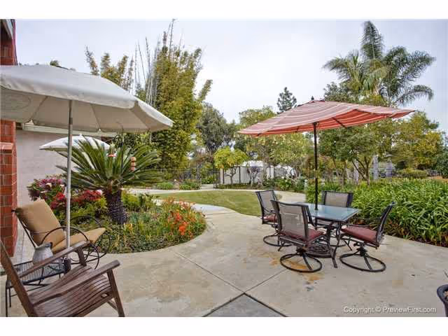 Outdoor patio area with a concrete walkway surrounded by lush greenery and flowering plants. There are two patio umbrellas, one white and one red, shading a glass-top table with four chairs. Additional seating includes a wooden chair with cushions. Trees and shrubs are visible in the background under a cloudy sky.