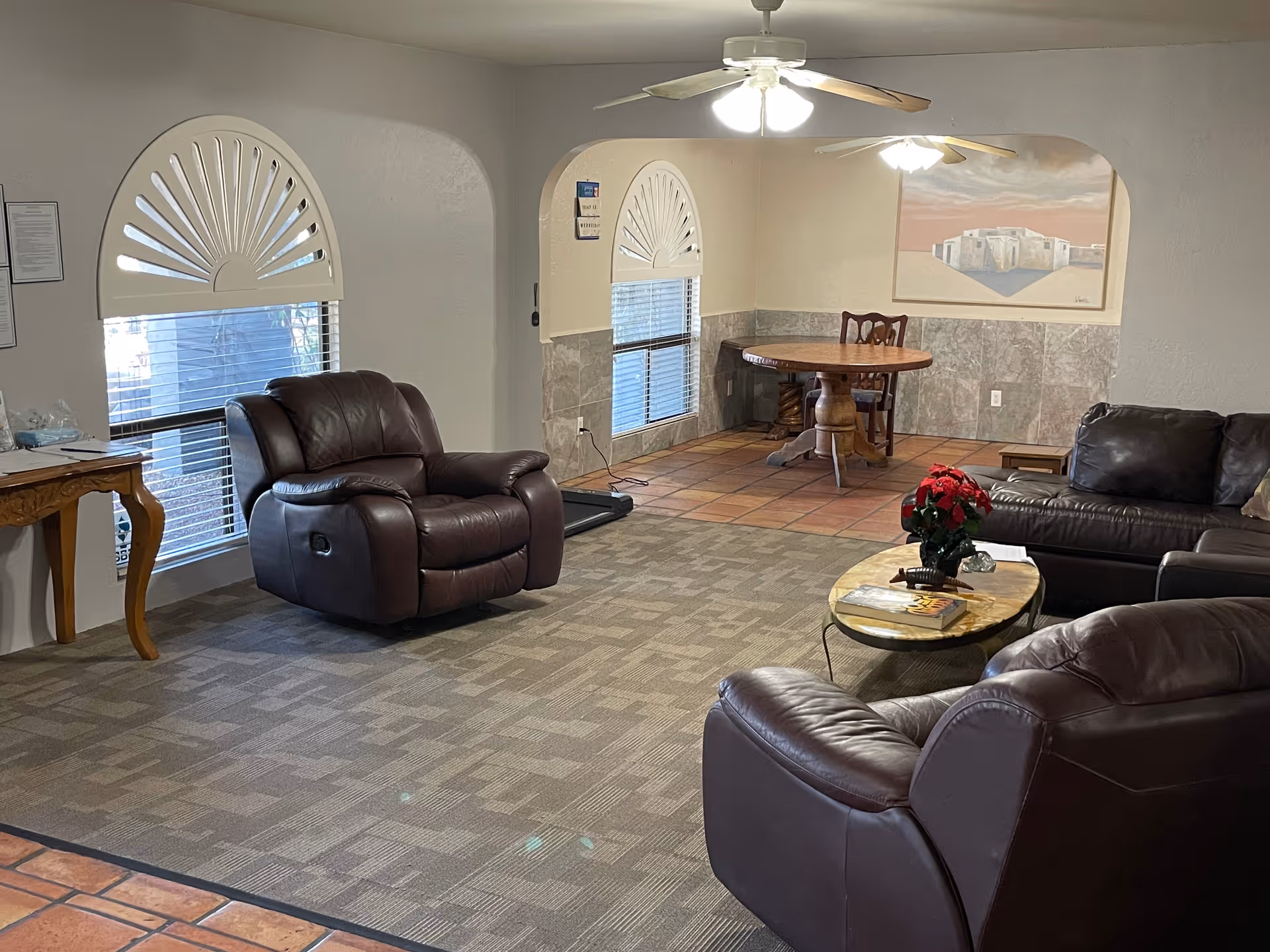 A cozy living room area with brown leather recliners and a sofa arranged around a small oval coffee table with a red poinsettia plant on it. The room has carpeted flooring with a tiled section in the back where a round wooden table and chair are placed. Two windows with decorative white shutters allow natural light into the room. Ceiling fans with lights are mounted on the ceiling, and a painting hangs on the wall above the tiled area.