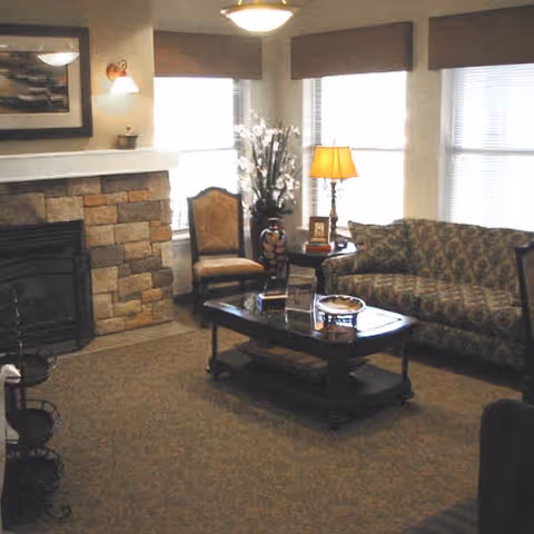 A cozy living room with a stone fireplace on the left, a patterned sofa on the right, a wooden coffee table in the center with decorative items, a chair near the window, a table lamp, and a vase with flowers by the window.