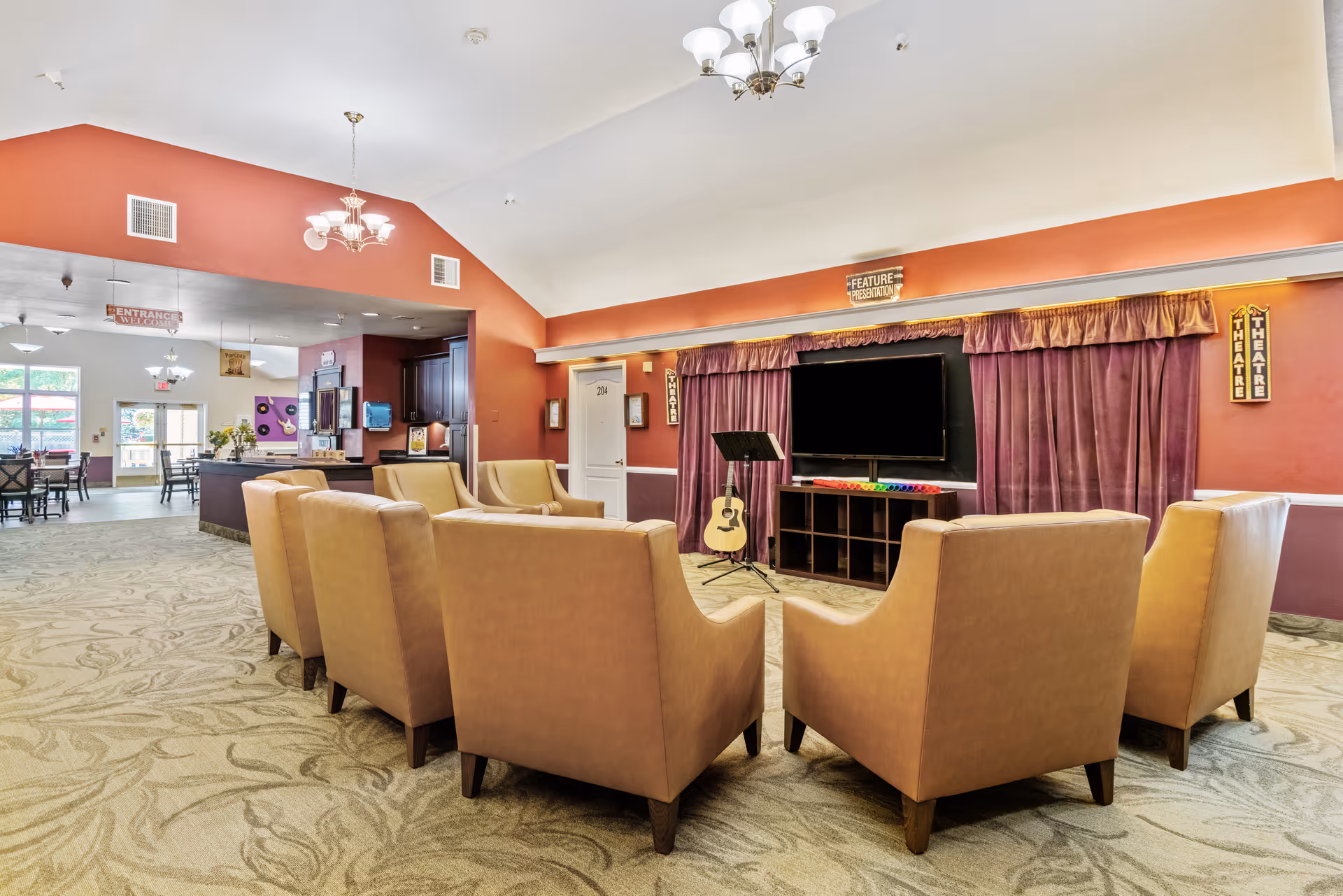 A spacious common area in a senior living facility with beige armchairs arranged in a semi-circle facing a large flat-screen TV mounted on a stand. Behind the TV are purple curtains and signs that say 'THEATRE' and 'FEATURE PRESENTATION'. The room has a high ceiling with two chandeliers, red and white walls, and patterned carpet. In the background, there is a dining area with tables and chairs and a reception or service counter.