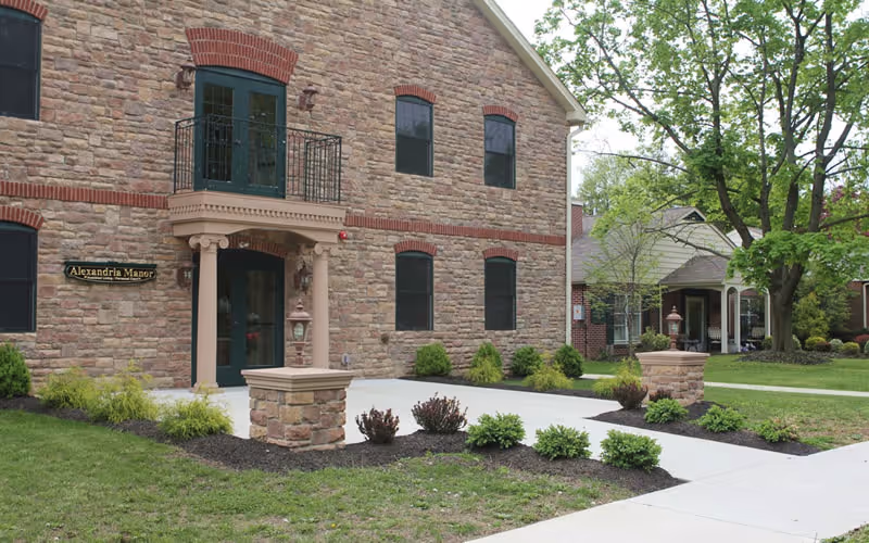 Exterior view of a two-story stone building with a small balcony above the entrance. The building has multiple windows with red brick accents and a sign that reads Alexandria Manor. There are landscaped bushes and a tree near the sidewalk leading to the entrance.
