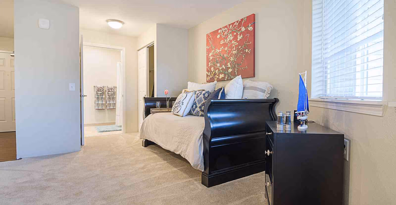 A bright bedroom with a black wooden daybed adorned with multiple decorative pillows. A red floral painting hangs above the bed. To the right, there is a black nightstand with a small blue sailboat model and other decorative items. A window with white blinds allows natural light into the room. In the background, an open door reveals a bathroom with two patterned towels hanging on the wall.