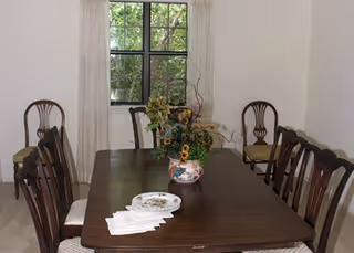 A dining room with a long wooden table surrounded by wooden chairs with cushioned seats. On the table, there is a decorative floral arrangement in a vase and a stack of white napkins with a small plate on top. A window with white curtains is visible in the background, letting in natural light.