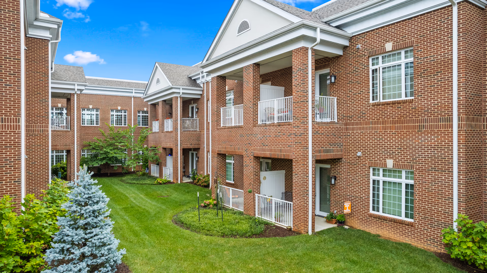 Exterior view of a two-story brick building with white trim, featuring balconies and large windows overlooking a well-maintained green lawn with shrubs and small trees under a blue sky with a few clouds.