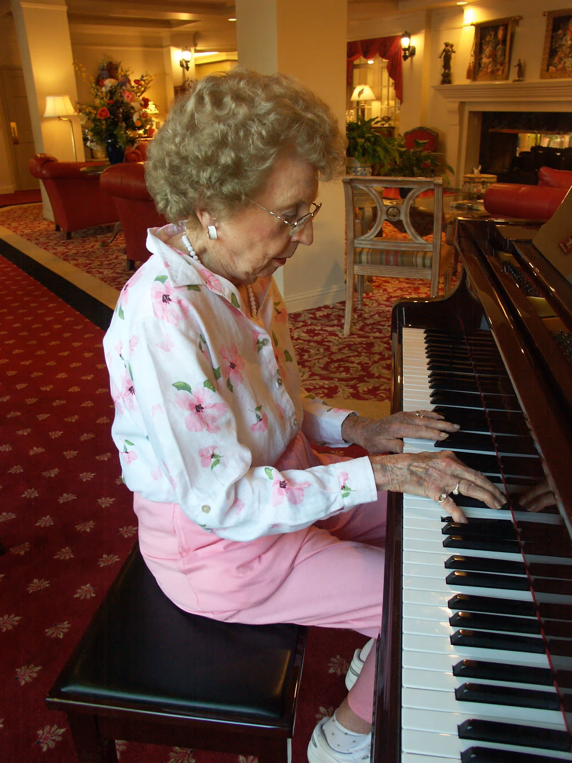 An elderly woman with curly gray hair, wearing glasses, a white blouse with pink floral patterns, and pink pants, is seated on a black piano bench playing a grand piano in a warmly lit room with red patterned carpet and classic furniture.