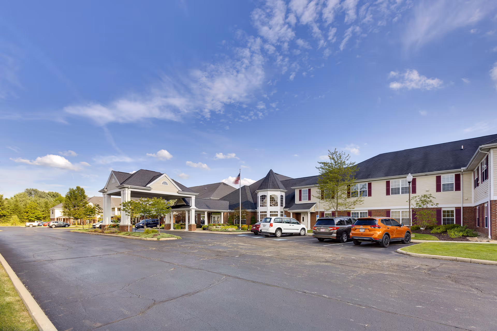Exterior view of Independence Village of Aurora, showing a large two-story building with a covered entrance, several parked cars in front, and a clear blue sky with some clouds.