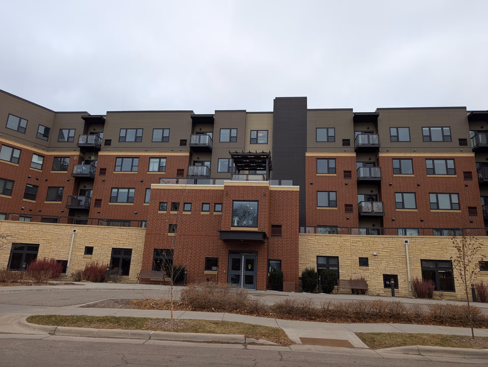 Exterior view of a multi-story senior living facility building with a mix of brick and stone facade, multiple windows, balconies, and a central entrance under an overcast sky.