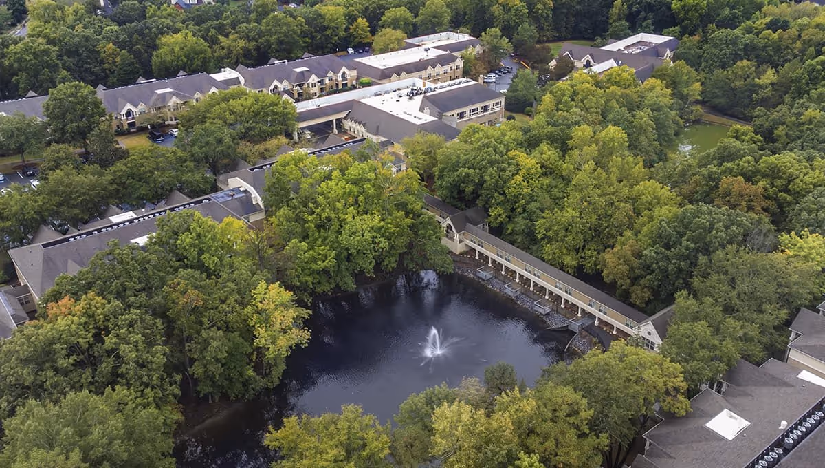 Aerial view of Brookdale Carriage Club Providence senior living facility surrounded by dense green trees with a central pond featuring a water fountain.