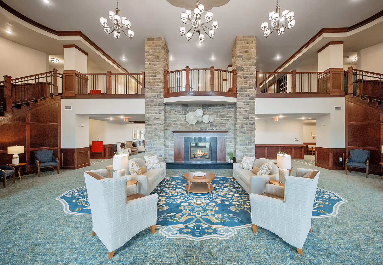 Spacious senior living facility common area with a large stone fireplace centered between two stone pillars. The room features two beige sofas and two light blue armchairs arranged around a wooden coffee table on a blue patterned rug. The area has high ceilings with three chandeliers, wood paneling, and a balcony railing above the fireplace.