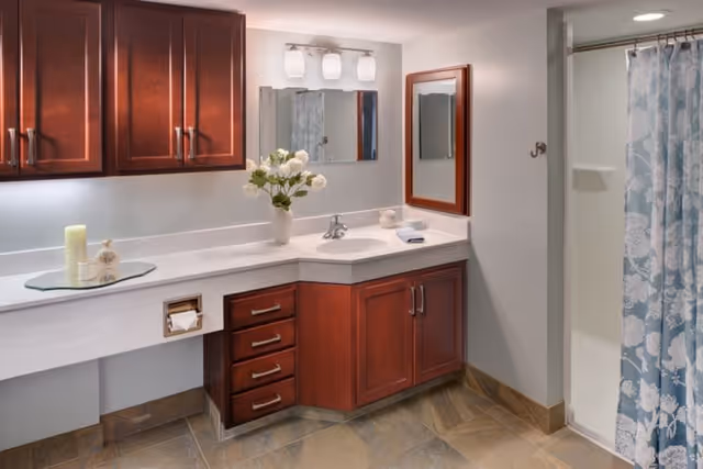 A clean and well-lit bathroom featuring a white countertop with a built-in sink, wooden cabinets and drawers with silver handles, a wall-mounted mirror with a wooden frame, a three-light fixture above the mirror, a vase with white flowers on the counter, a soap dispenser, a towel, and a shower area with a blue and white floral shower curtain.