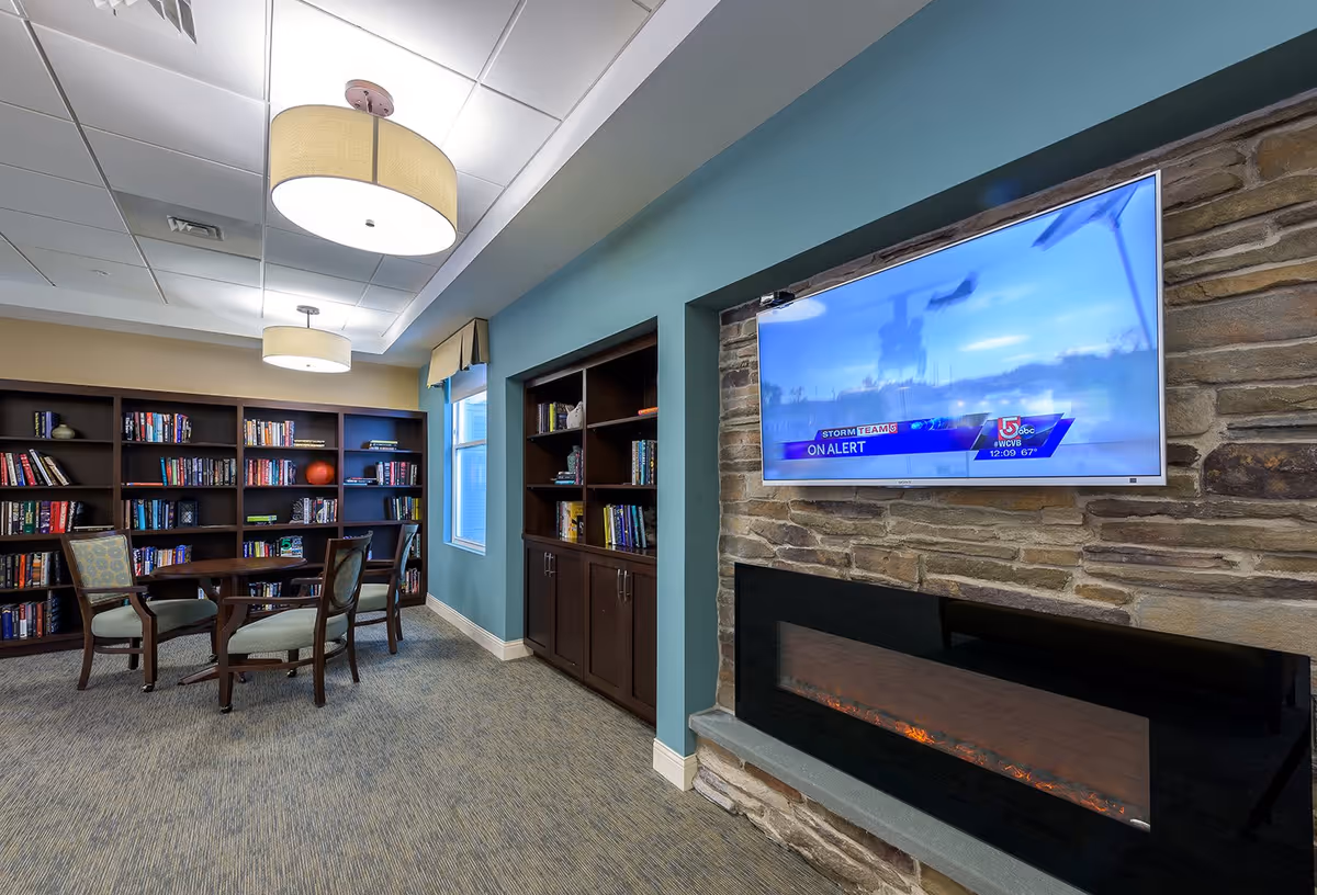 A cozy common area with bookshelves filled with books, a round table with four chairs, two large ceiling lights, a window with a valance, a stone wall with a mounted flat-screen TV displaying a news channel, and an electric fireplace below the TV.