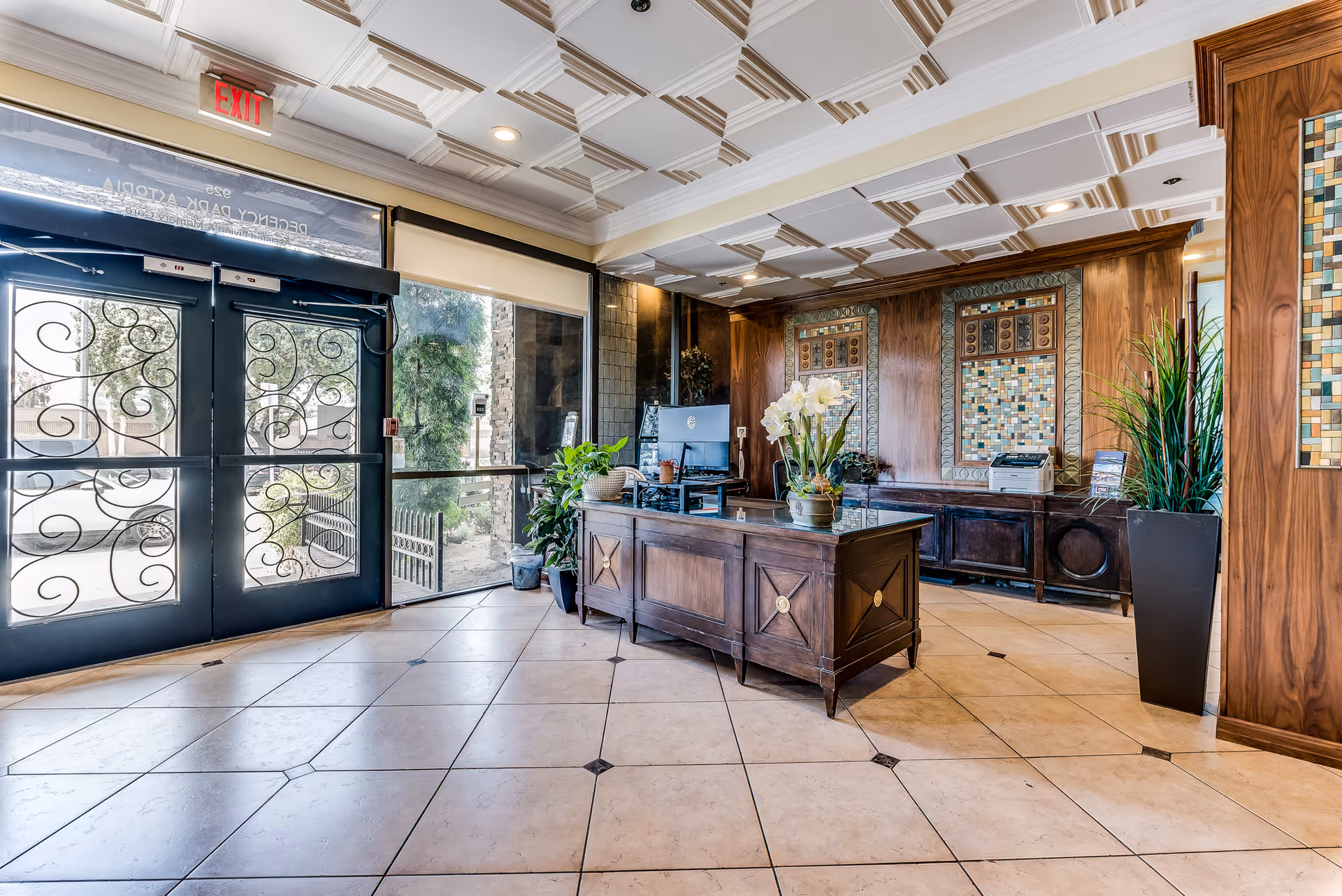 Reception area of Astoria Park Senior Living featuring a wooden front desk with a computer and decorative plants. The room has tiled flooring, a coffered ceiling with recessed lighting, and decorative mosaic wall panels. Large glass doors with ornate metal designs lead outside, and an exit sign is visible above the doors.