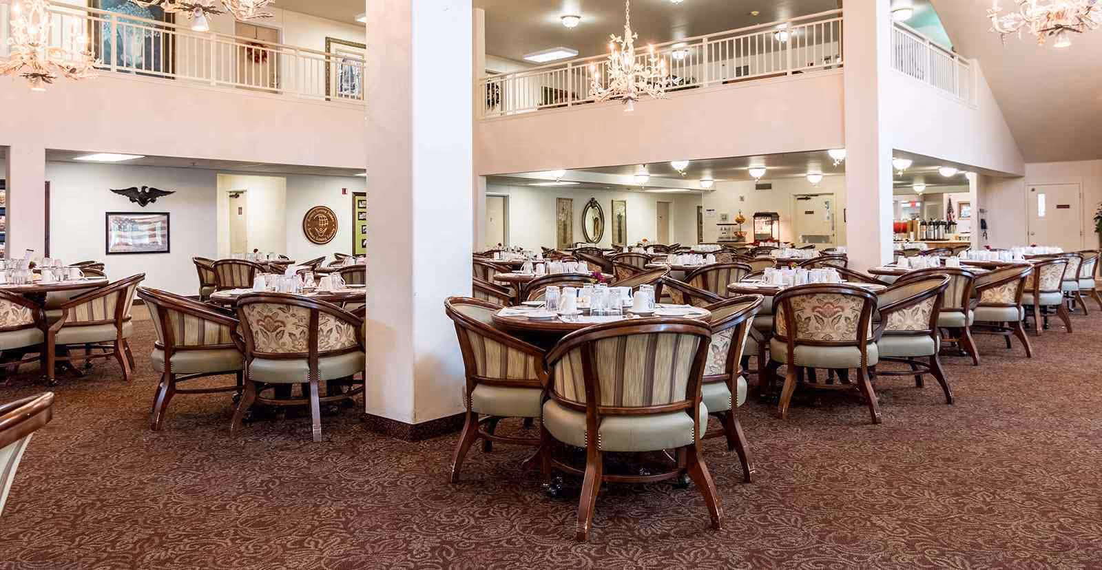 A spacious dining room with multiple round tables set with white cups, saucers, and silverware. Each table is surrounded by cushioned wooden chairs with patterned upholstery. The room features a high ceiling with chandeliers and a second-floor balcony railing overlooking the dining area. The carpet has a detailed pattern, and the walls are decorated with framed artwork and mirrors.