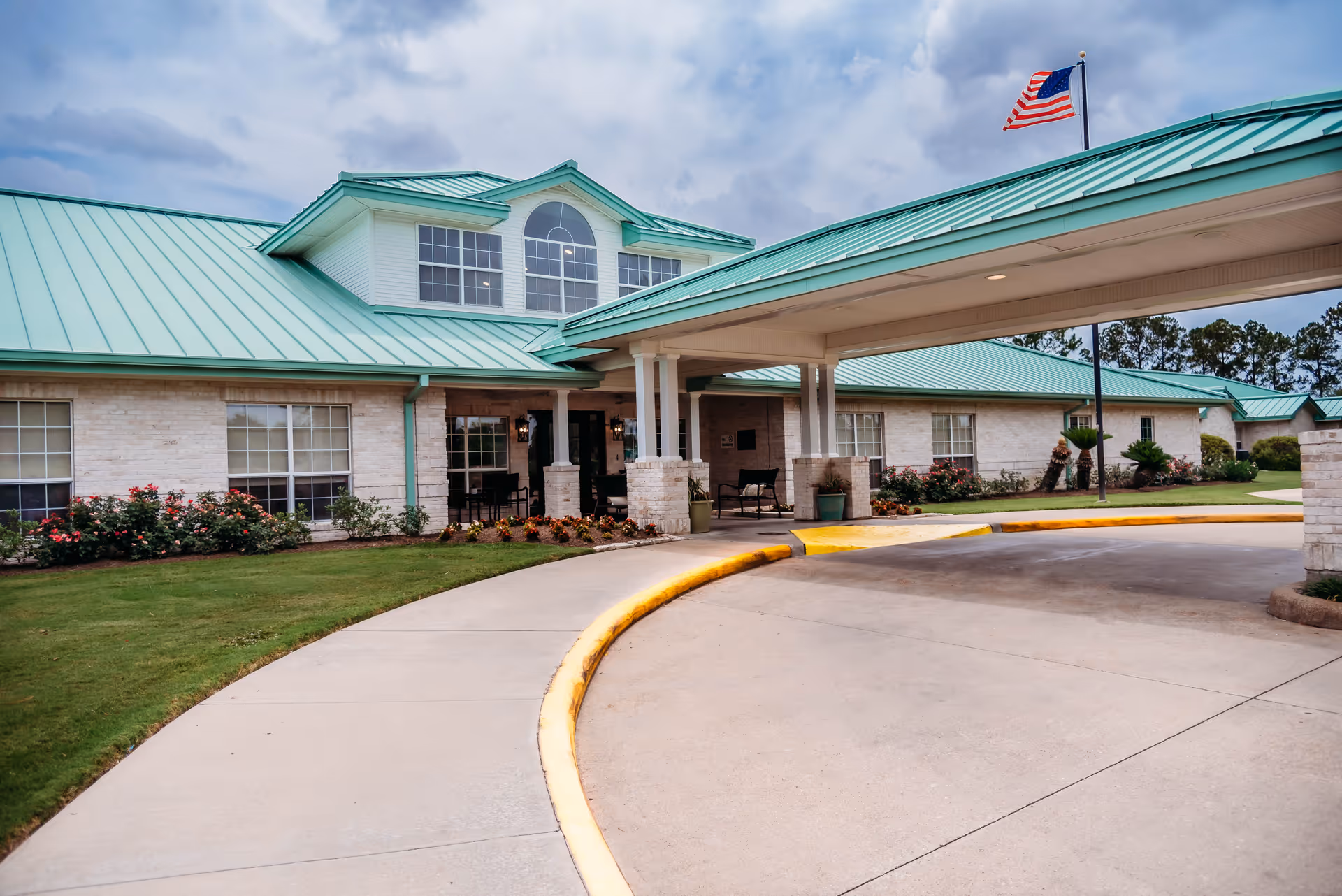 Exterior view of a single-story nursing and rehabilitation center building with a green metal roof, large windows, and a covered entrance driveway. There is a well-maintained lawn with flower beds and an American flag flying on a flagpole near the entrance.