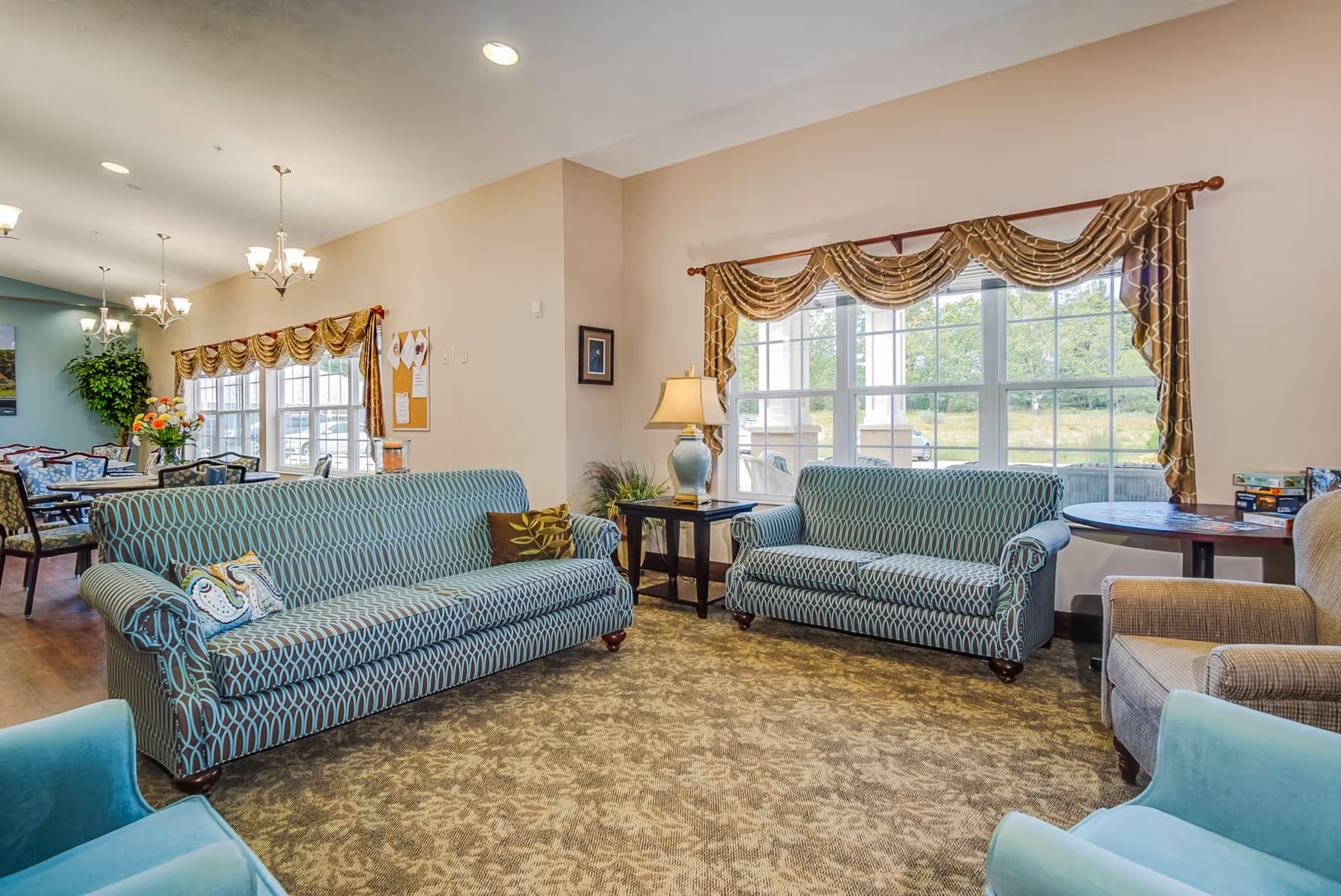A bright and spacious living room area in an assisted living facility featuring two patterned blue sofas, two light blue armchairs, a beige armchair, a side table with a lamp, and large windows with decorative curtains letting in natural light. In the background, there is a dining area with tables, chairs, and hanging light fixtures.