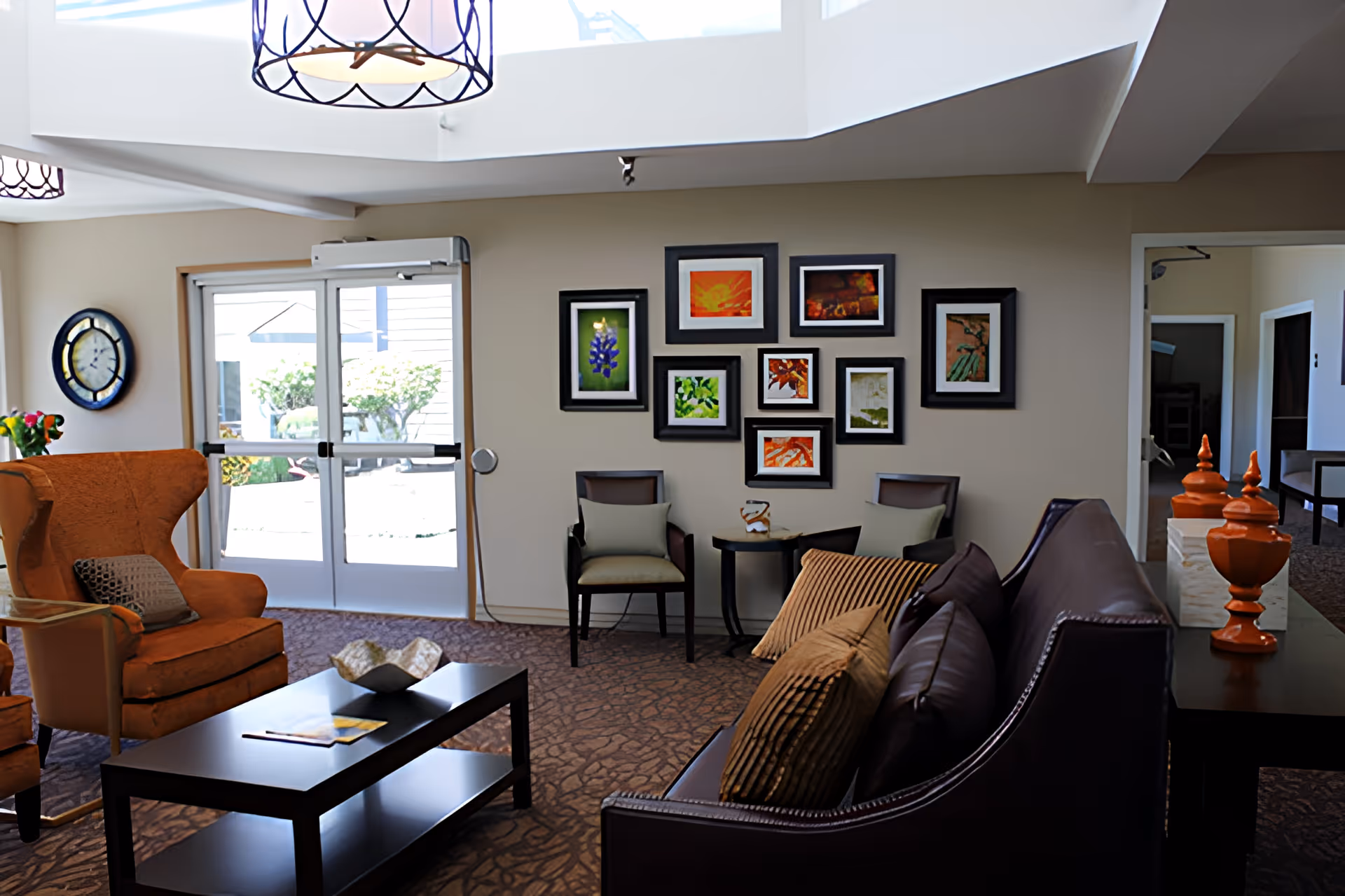 A cozy senior living facility common area with a brown leather sofa adorned with striped pillows, an orange armchair, two chairs with cushions, a black coffee table with decorative items, and a wall decorated with multiple framed colorful pictures. There is a large clock on the wall near a glass door leading outside, and two hanging light fixtures above.