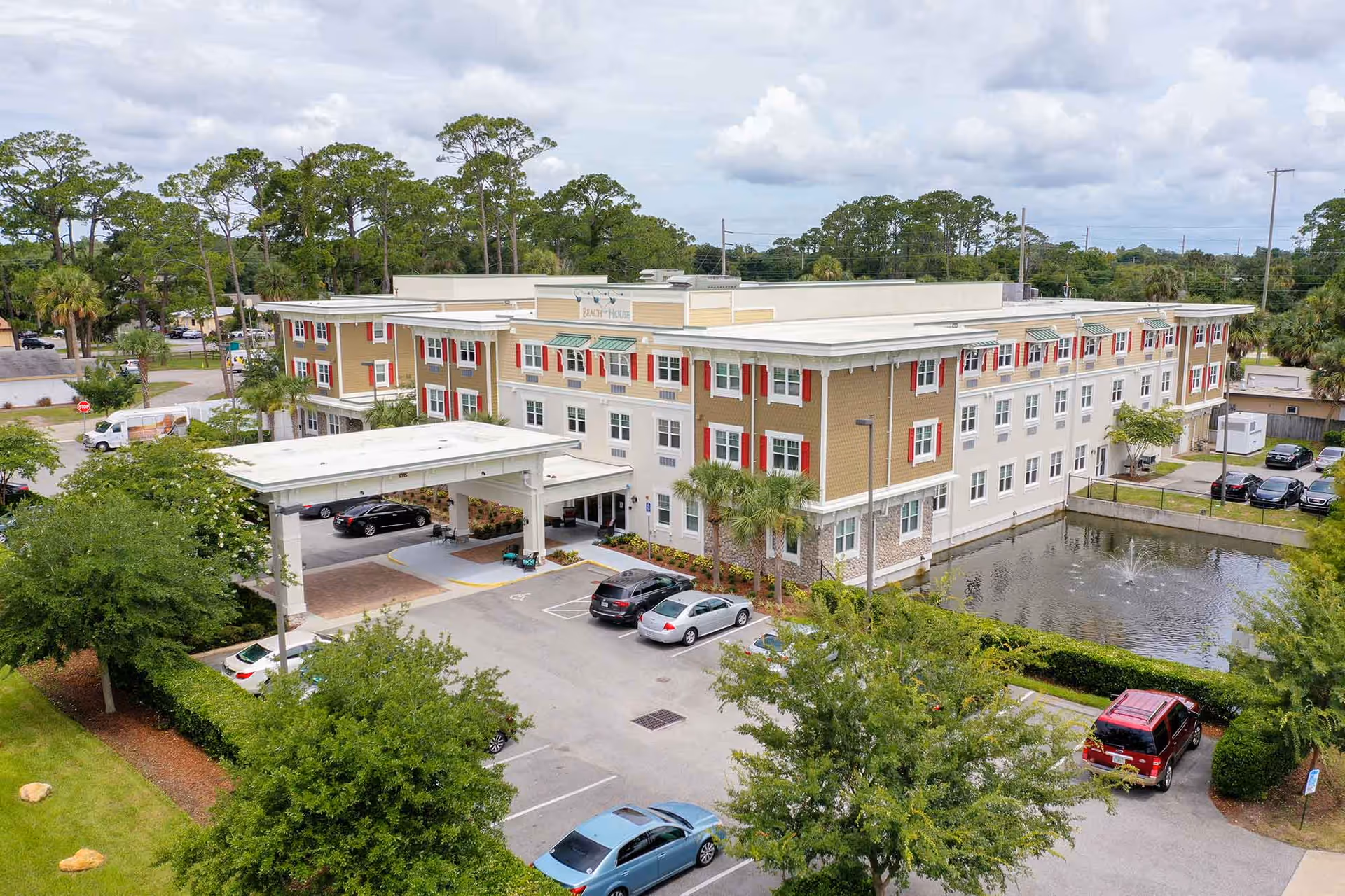 Aerial view of Beach House Assisted Living & Memory Care building with a covered entrance, parking lot with several cars, a small pond with a fountain, and surrounding trees and greenery under a partly cloudy sky.