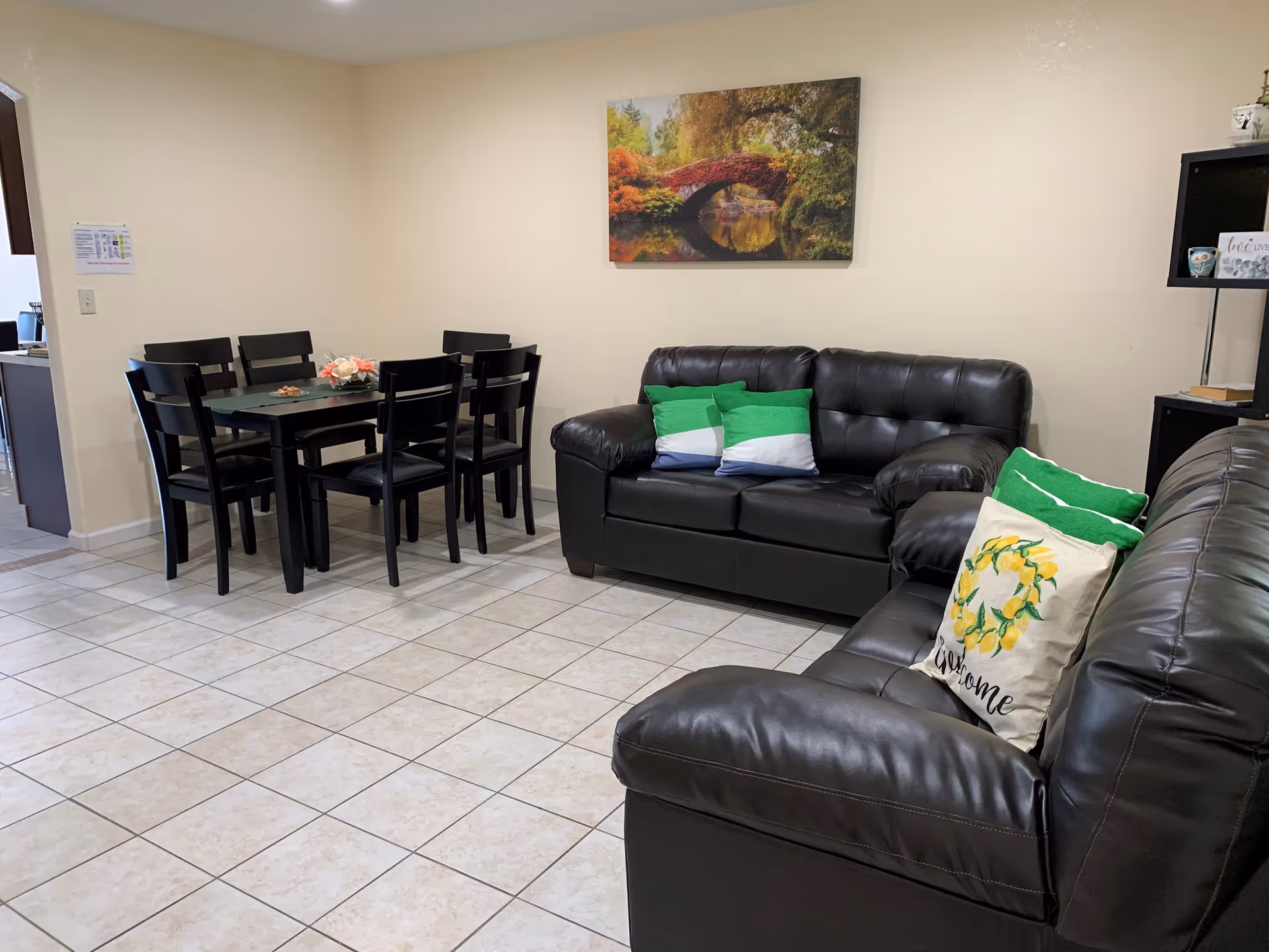 A cozy living room area with two black leather sofas adorned with green and white pillows, one pillow featuring a lemon design and the word 'welcome'. A black dining table with six matching chairs is positioned against a beige wall. Above one sofa hangs a colorful painting of a stone bridge over a calm river surrounded by autumn trees. The floor is tiled with light-colored tiles, and a black shelving unit with decorative items is visible on the right side.