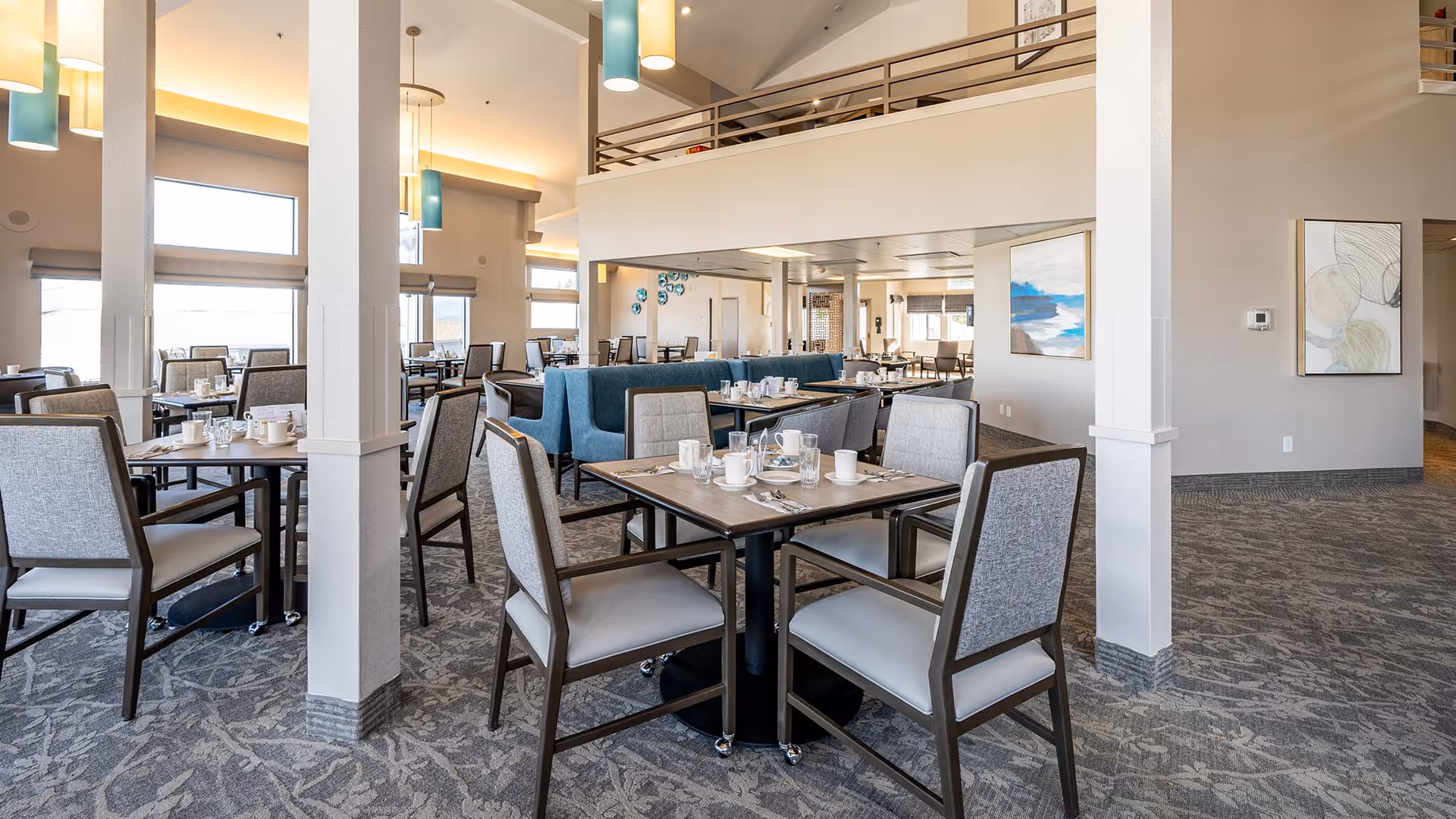 A spacious and well-lit dining room with multiple tables and chairs arranged neatly. The room features high ceilings with modern pendant lights, large windows allowing natural light to fill the space, and contemporary artwork on the walls. Tables are set with cups, glasses, and utensils, ready for dining.