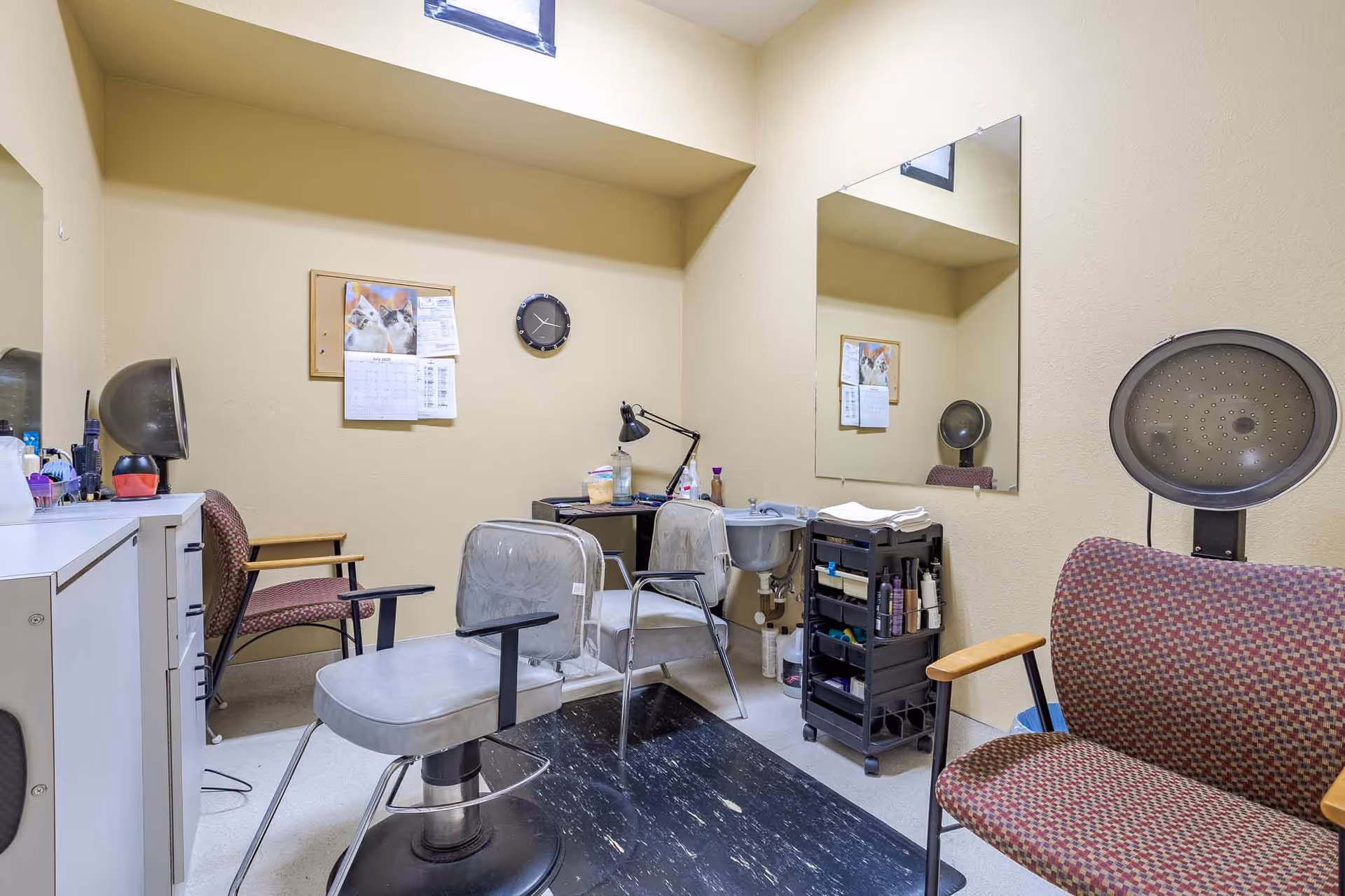 A small salon room with two salon chairs, a hair dryer chair, a large mirror on the wall, a rolling cart with hair products, a small sink, a clock, and a bulletin board with papers pinned on it.