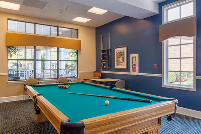 A recreational room with a pool table in the center, two pool cues and a white cue ball on the table. The room has large windows with yellow and beige valances, blue and beige walls, framed artwork, and a rack holding additional pool cues. Outside the windows, greenery and a patio area are visible.