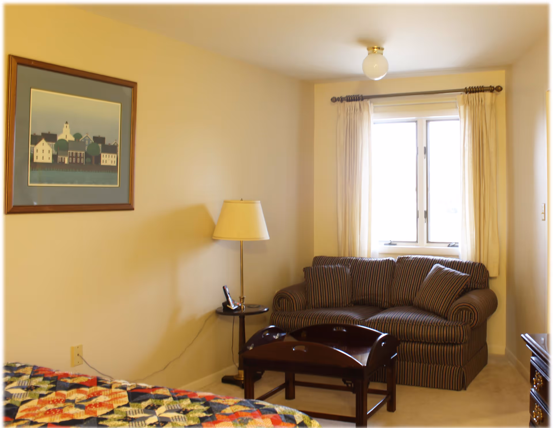A cozy room with a striped loveseat under a window with cream-colored curtains. There is a wooden side table with a lamp and a cordless phone next to the loveseat. A colorful quilt is partially visible on a bed in the foreground, and a framed picture of houses hangs on the wall.