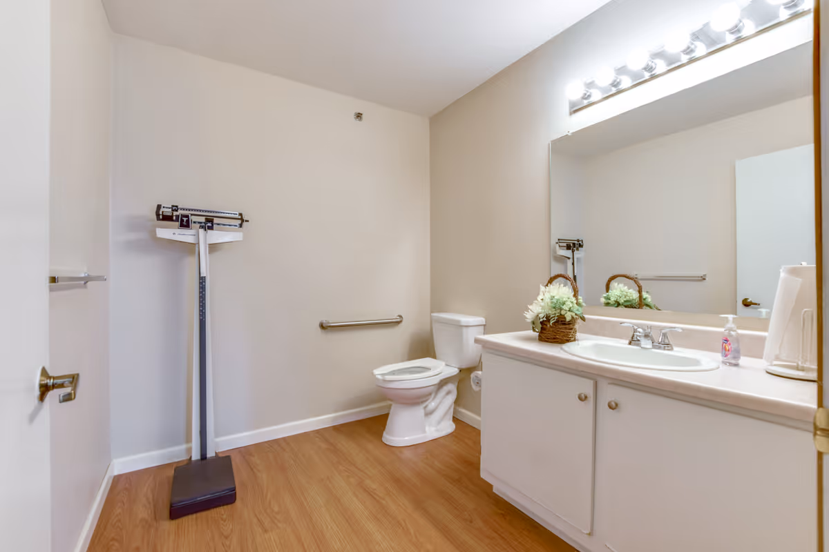 A clean bathroom with a white toilet, a large mirror above a white sink with countertop, a basket with white flowers on the counter, a paper towel holder, and a standing weight scale against the wall. The floor is wood-style laminate and the walls are light beige.