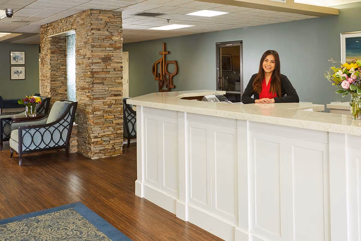 A smiling woman stands behind a white reception desk in a senior living facility. The room features wooden flooring, a stone pillar, comfortable seating with armchairs, floral arrangements, and wall art. The atmosphere is welcoming and well-lit.