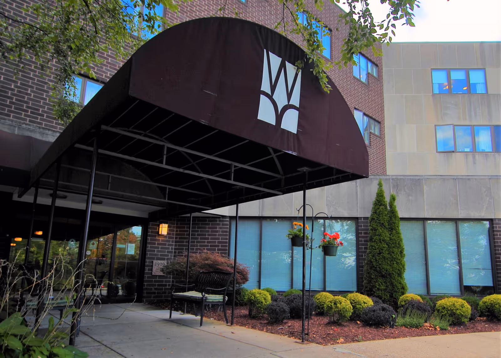 Entrance to a senior living facility with a large black canopy featuring a white logo. The building exterior is made of brick and stone with several windows. There is a bench near the entrance and landscaped bushes and hanging flower pots along the walkway.
