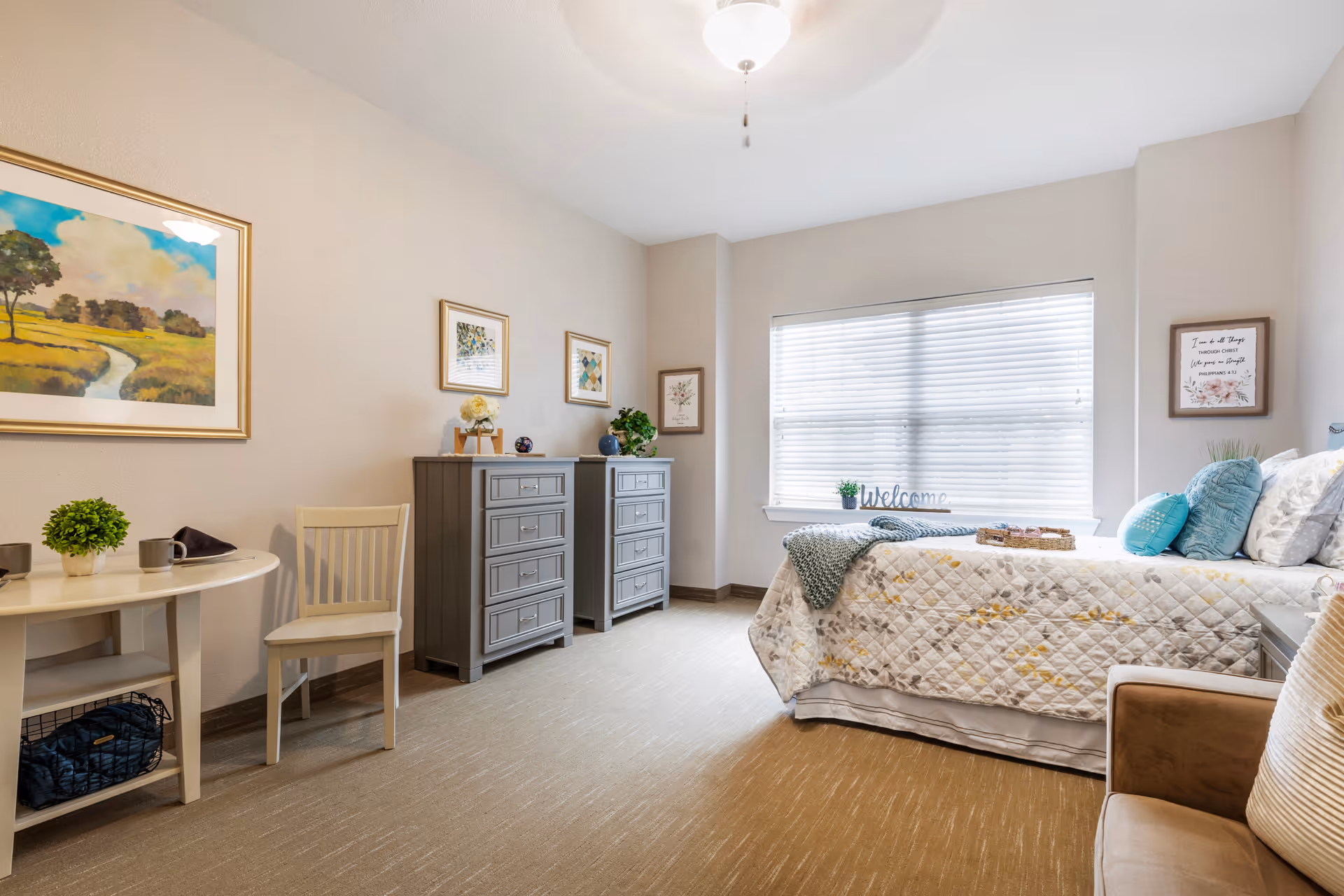 A bright, furnished bedroom with a made bed by a large window, two gray dressers, a small table and chair, and decorative wall art.