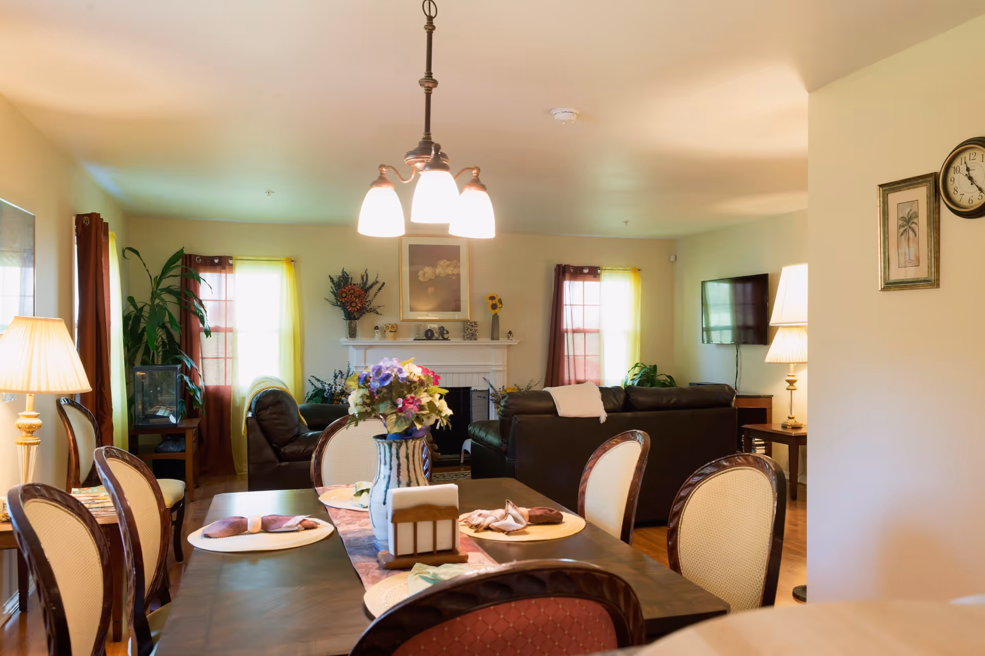 A well-lit living and dining area in a senior living facility named Sam Sunflower. The dining table is set with placemats, napkins, and a vase of flowers. In the background, there are leather sofas, a fireplace with decorative items, a wall-mounted TV, and windows with curtains allowing natural light to enter. Lamps and framed artwork adorn the walls.