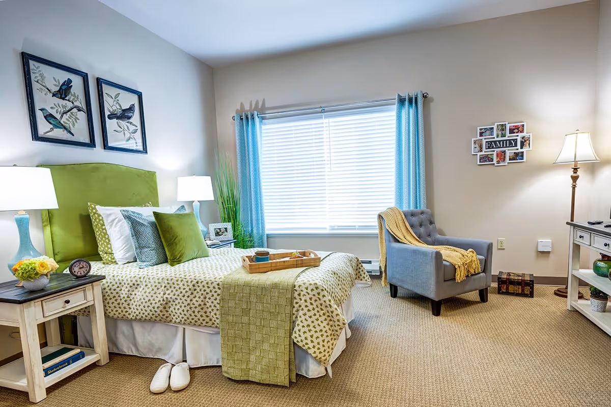 A cozy bedroom with a green upholstered headboard, a bed with green and white patterned bedding, and multiple pillows. There are two bedside tables with lamps, a pair of white slippers on the carpeted floor, a blue armchair with a yellow throw blanket, a window with blue curtains, and a floor lamp. The walls are decorated with framed bird artwork and a family photo collage.