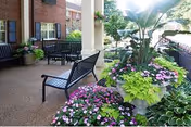 Outdoor patio area with black metal benches and a large planter filled with green foliage and colorful flowers, adjacent to a brick building with windows and surrounded by greenery.